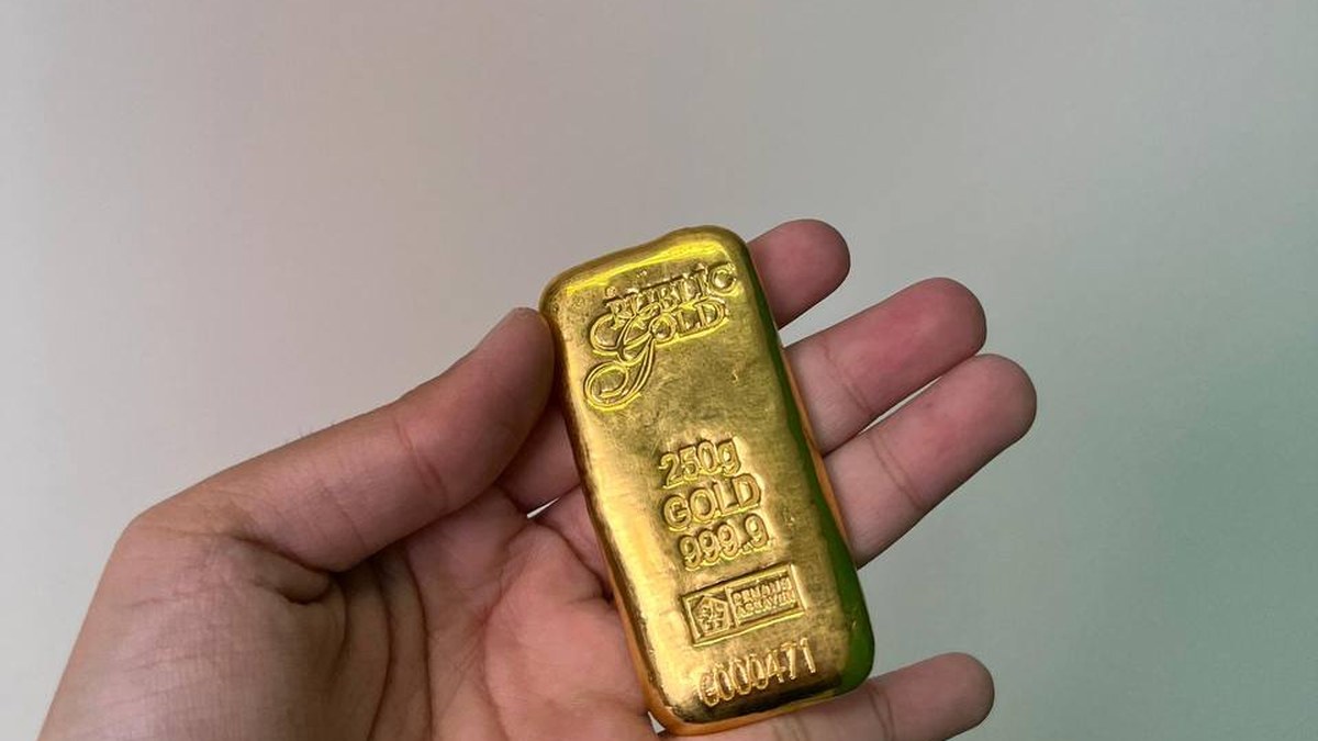 Tangan memegang emas batangan Public Gold 250 gram dengan latar dinding polos, ilustrasi modus penipuan emas berkedok kurir.